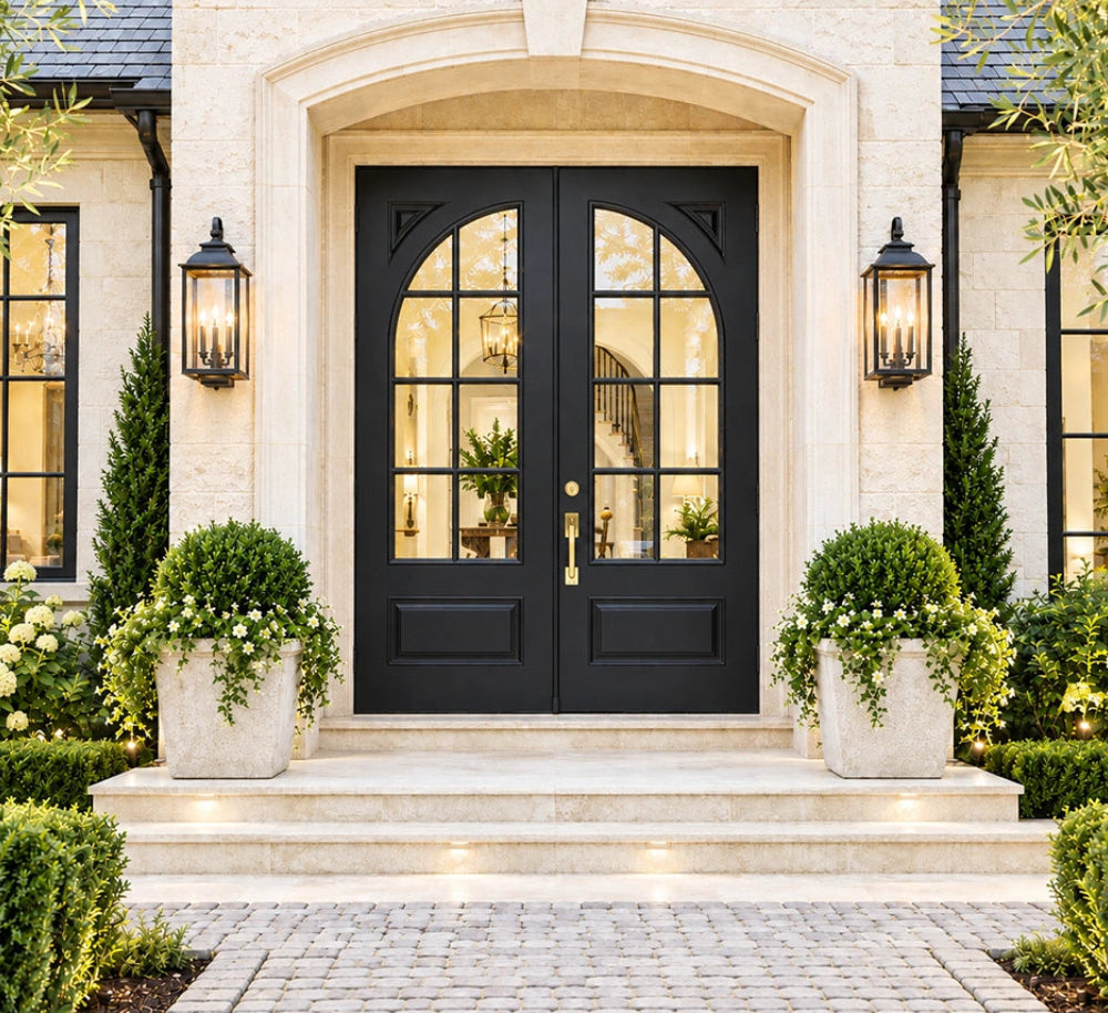 Black double front doors with arched top glass, set in a stone entryway with matching black wall sconces, planters, shrubs, and a paved walkway