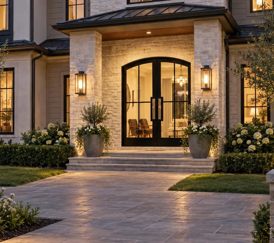 Modern house front entrance with black arched double doors, stone columns, warm wall lighting, and a paved walkway in a landscaped yard.